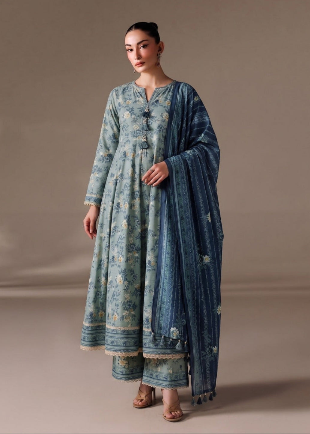A woman models a traditional three-piece outfit consisting of a light blue printed kurta, matching trousers, and a dark blue printed dupatta, set against a solid beige studio background.