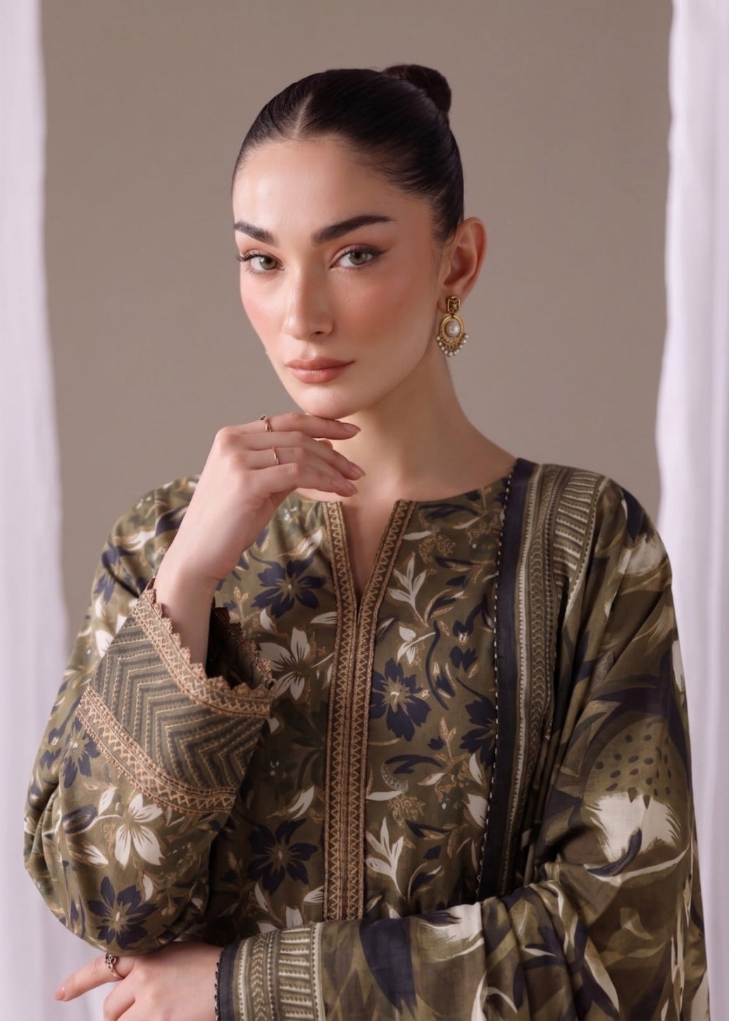A woman wearing a long-sleeve olive green printed tunic top with blue and beige floral motifs, styled with gold earrings against a neutral beige background.