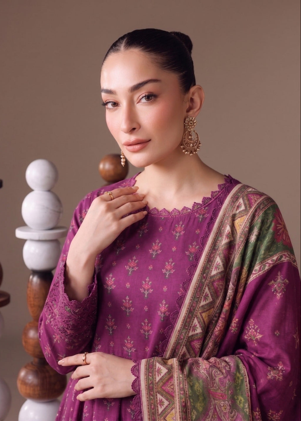 A woman modeling a traditional Indian outfit with a deep purple kurta featuring small floral prints, paired with a matching patterned dupatta, set against a warm beige background with decorative wooden and stone stacks.