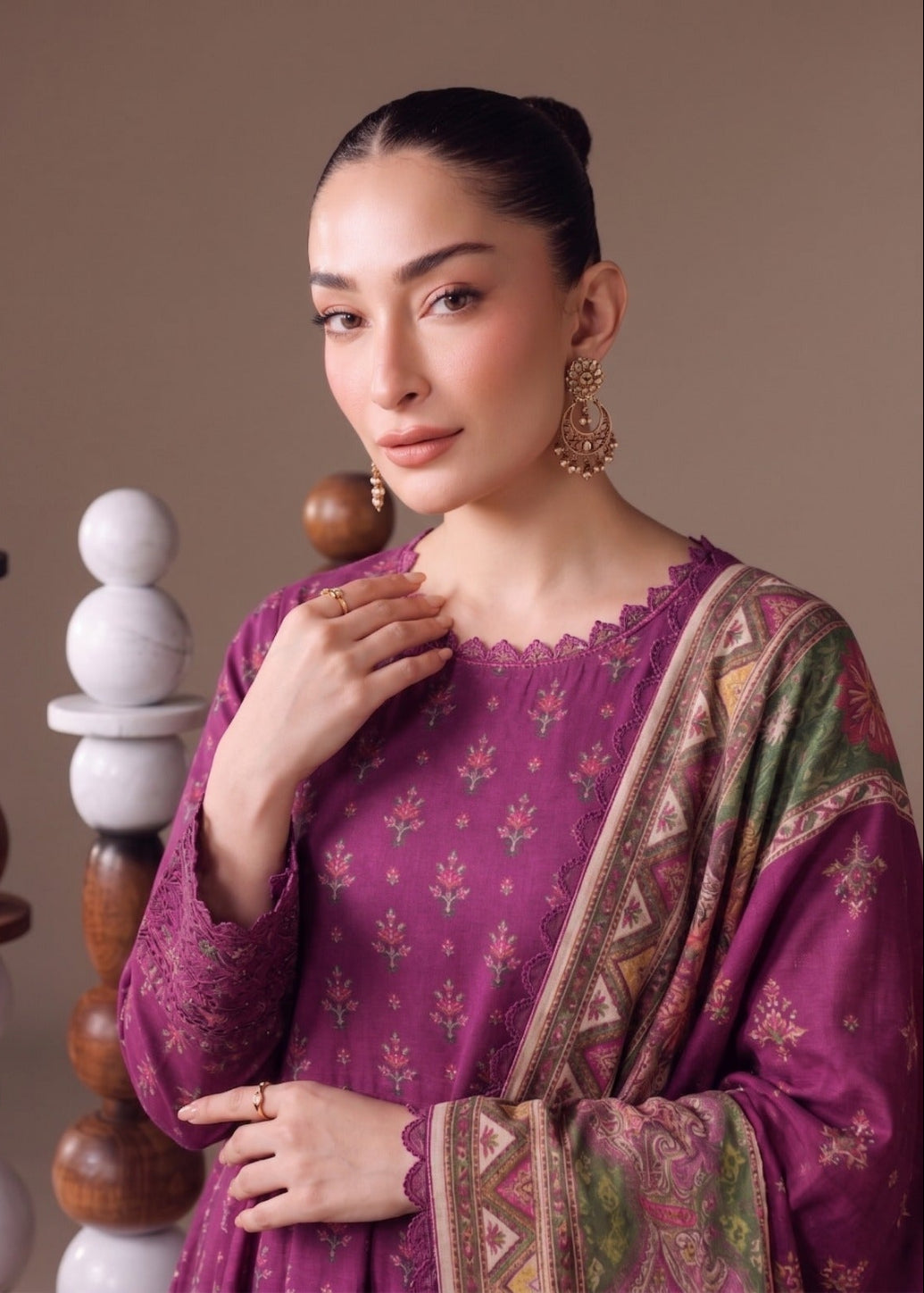 A woman modeling a traditional Indian outfit with a deep purple kurta featuring small floral prints, paired with a matching patterned dupatta, set against a warm beige background with decorative wooden and stone stacks.