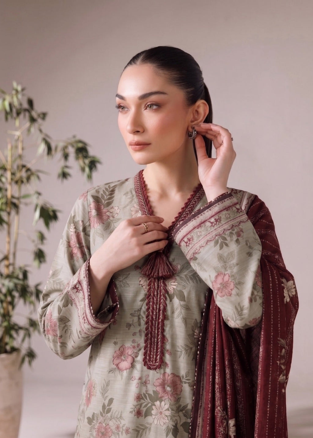 A woman models a traditional South Asian salwar kameez set featuring a sage green floral print with dusty pink and brown details, paired with a deep maroon embroidered dupatta and matching tassel tie.