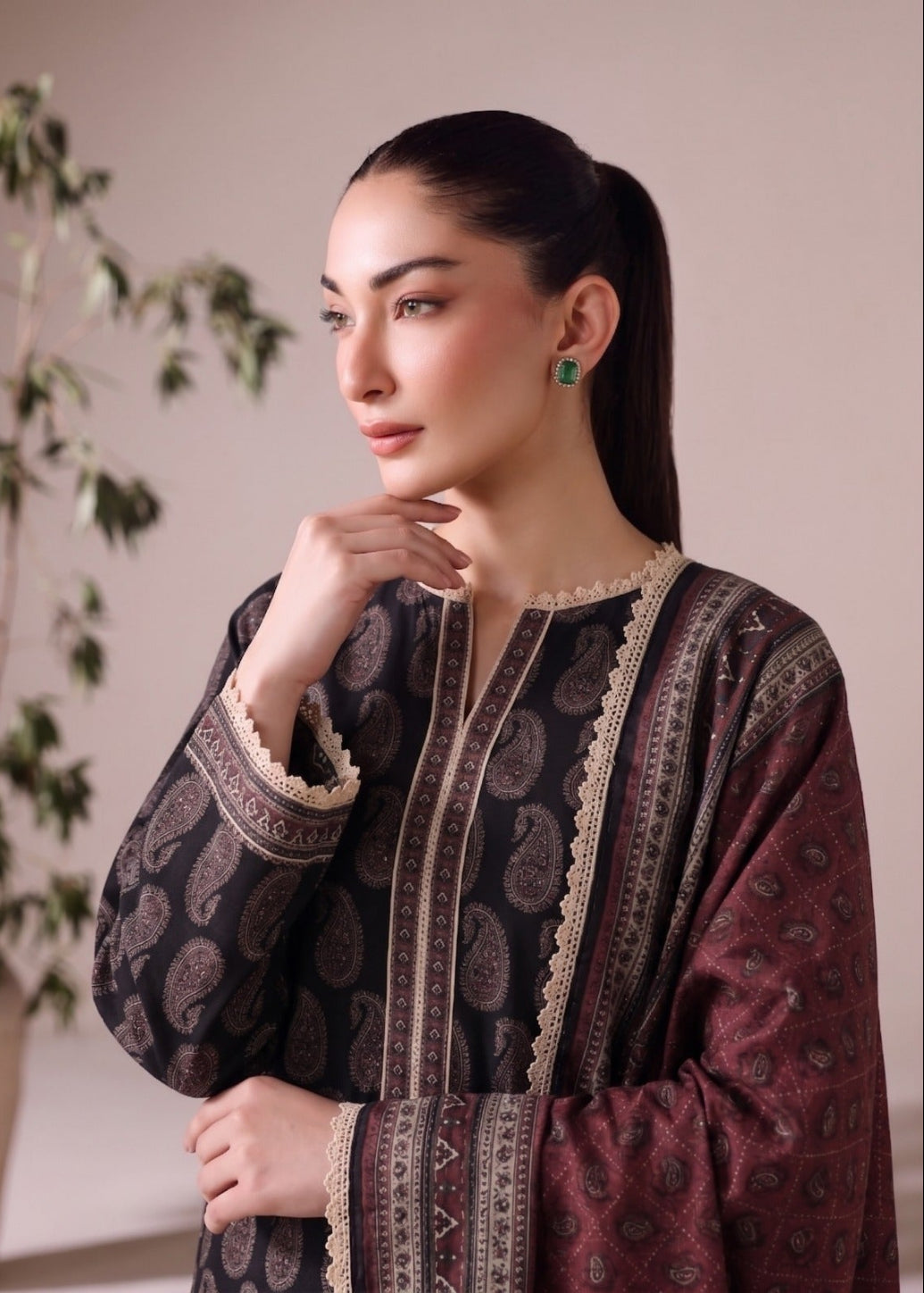 A woman wearing a dark brown and maroon patterned traditional long-sleeve kurta with beige lace trim, posing in an indoor setting with a soft beige background and a blurred plant.