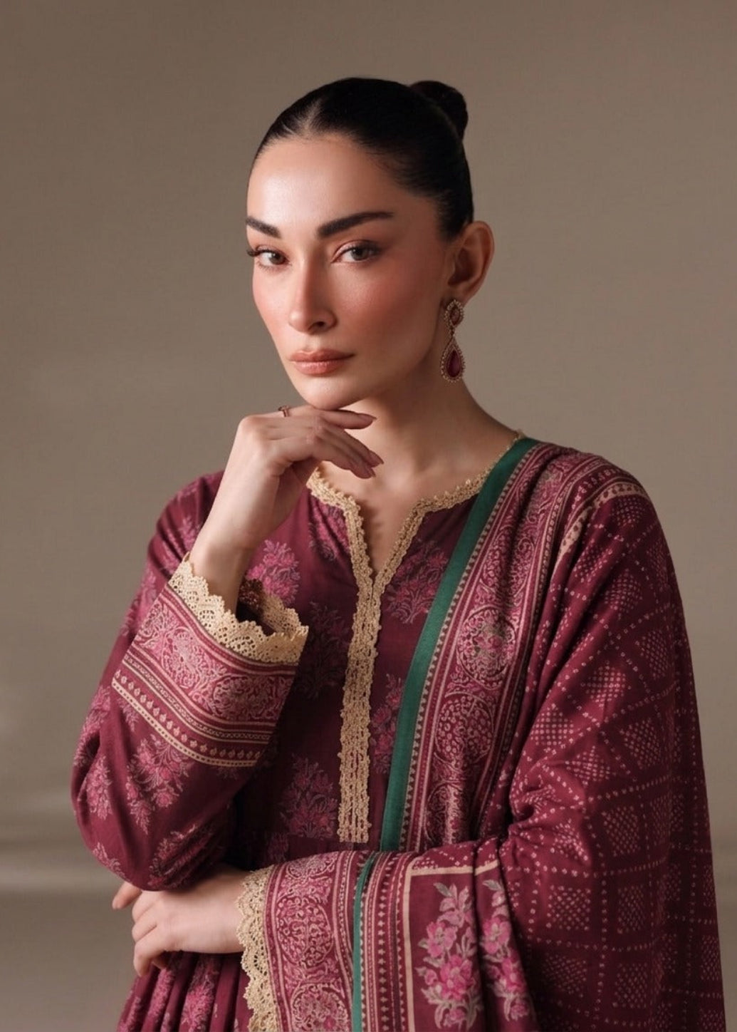 A woman wearing a traditional maroon embroidered kurta with beige lace trim and green border details, posed against a neutral beige background.