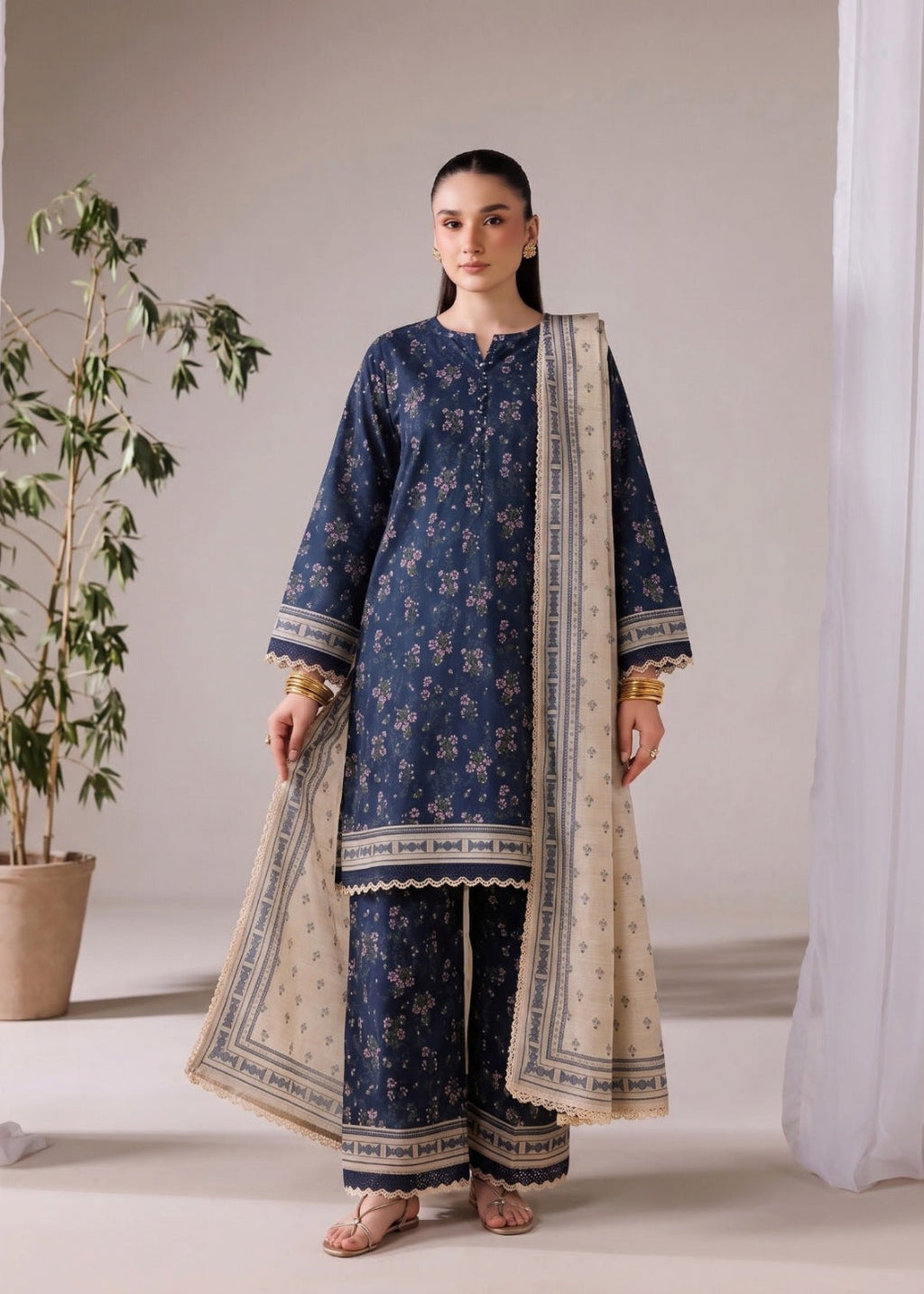 A woman modeling a navy blue floral printed three-piece traditional outfit with a beige dupatta, standing in a minimalist studio setting with a potted plant and white drapes.