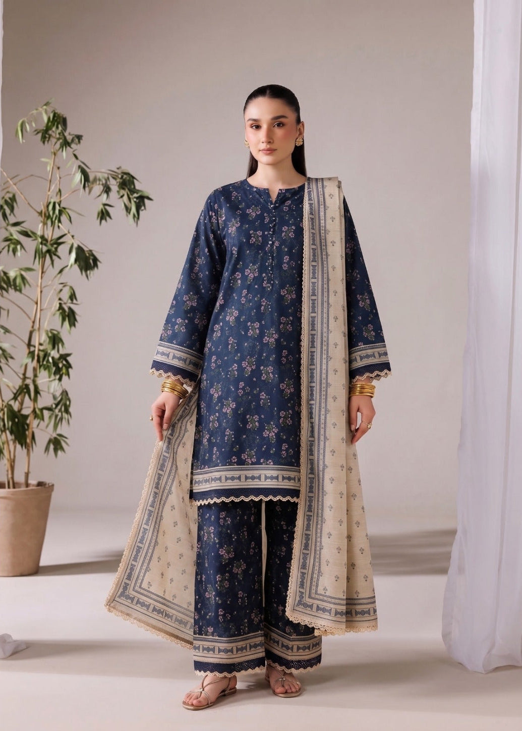 A woman modeling a navy blue floral printed three-piece traditional outfit with a beige dupatta, standing in a minimalist studio setting with a potted plant and white drapes.