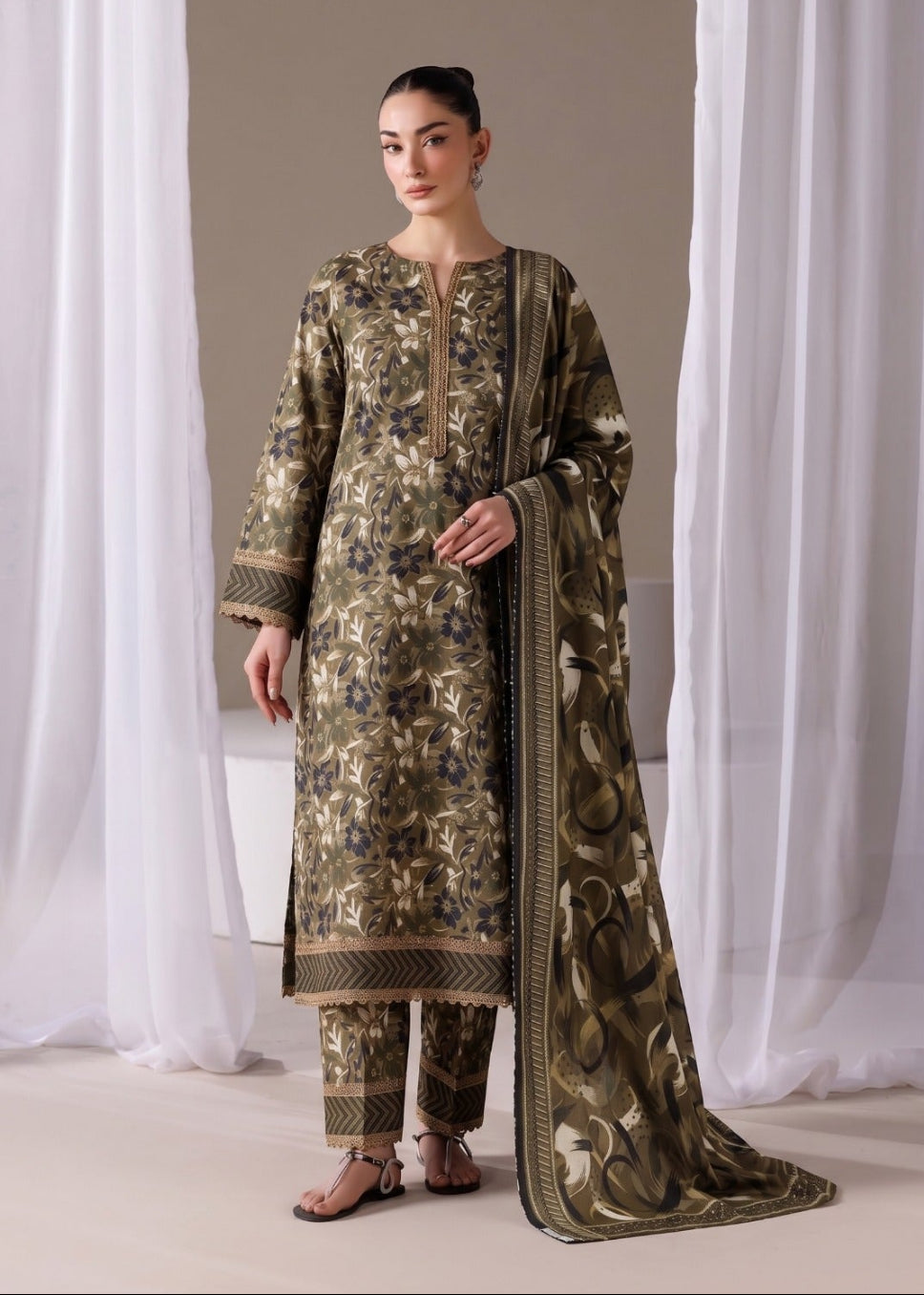 A woman models a three-piece traditional outfit with a matching dupatta. The set features an olive green base with intricate gold and blue floral embroidery, paired with matching trousers and a long draped dupatta with gold borders.