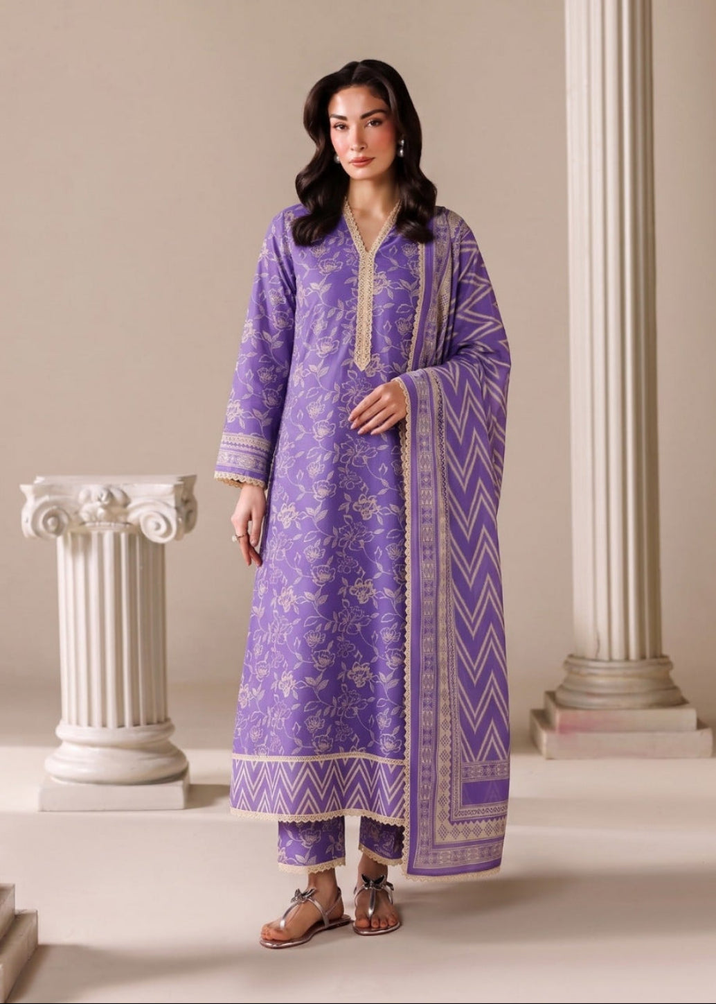 A woman modeling a traditional purple three-piece salwar kameez suit with beige/gold embroidery and a matching patterned dupatta, standing between classical columns in a beige studio setting.
