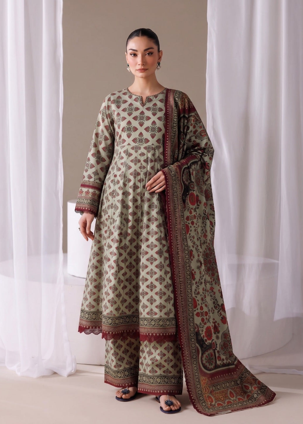A woman models a traditional three-piece salwar kameez suit set in a neutral studio setting with white sheer curtains. The set features an olive green kurta and wide-leg trousers with a beige and red geometric pattern, paired with a matching dupatta that has a red border and floral motifs.