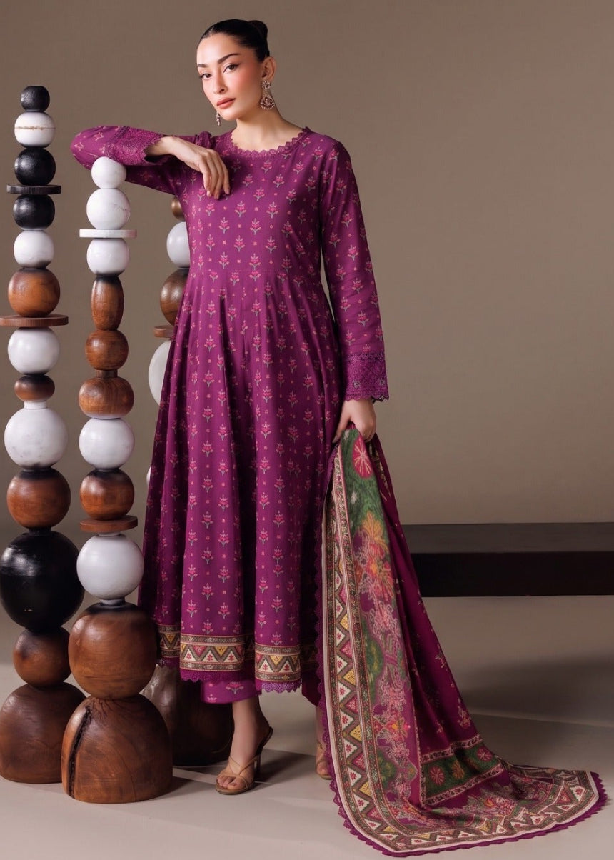 A woman models a traditional three-piece outfit consisting of a magenta-purple long kurta with gold floral prints, matching trousers, and a patterned dupatta, standing next to a decorative wooden and stone sculpture against a solid beige background.