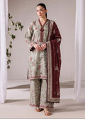 A woman modeling a traditional three-piece salwar kameez suit in sage green with pink floral prints and a deep maroon dupatta with gold borders, set against a neutral studio backdrop with sheer white curtains.