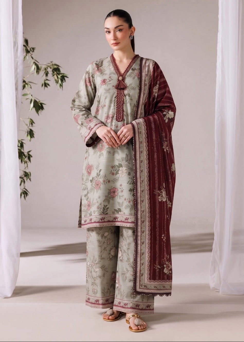 A woman modeling a traditional three-piece salwar kameez suit in sage green with pink floral prints and a deep maroon dupatta with gold borders, set against a neutral studio backdrop with sheer white curtains.