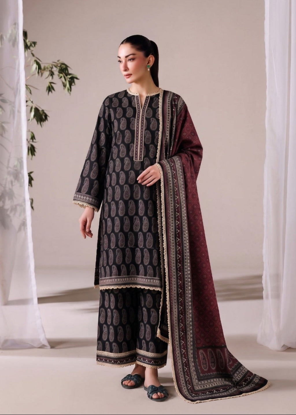A woman models a traditional three-piece salwar kameez suit in a studio setting. The set features a dark gray printed kurta and matching trousers with gold borders, paired with a deep maroon dupatta that has gold and silver patterns.