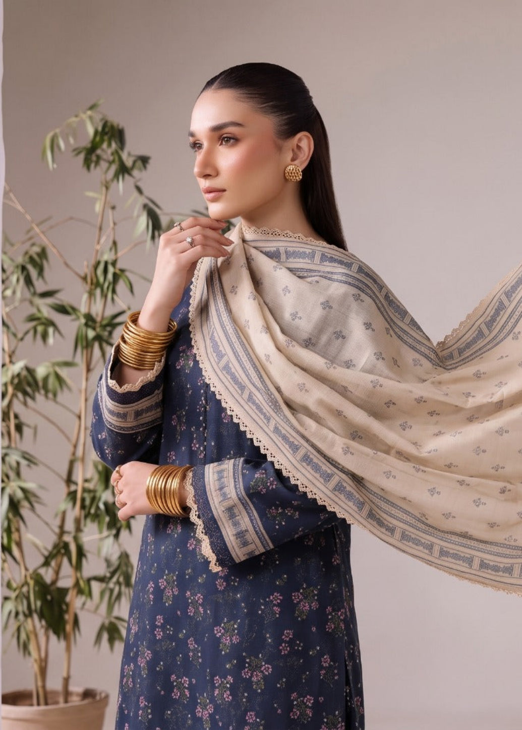 A woman wearing a navy floral traditional outfit holds up a large cream-colored dupatta with a blue border and small geometric motifs, set against a neutral indoor background with a potted plant.