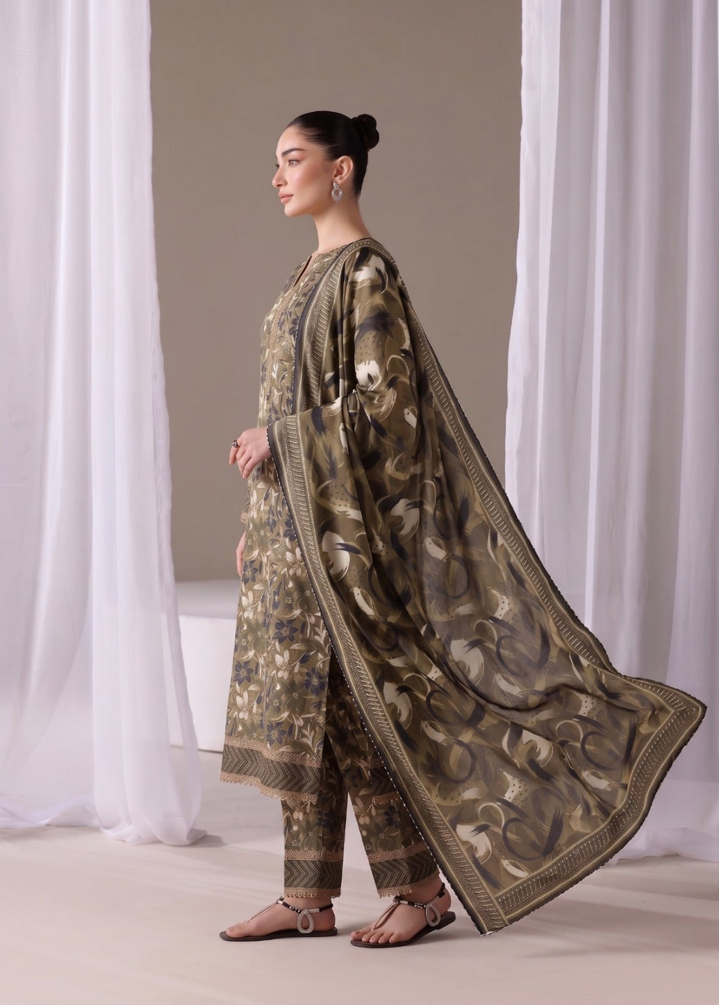 A woman models a traditional three-piece salwar kameez suit in olive green and beige with intricate floral prints, featuring a matching sheer dupatta with a printed paisley pattern and gold borders, set against a neutral studio backdrop with white drapes.