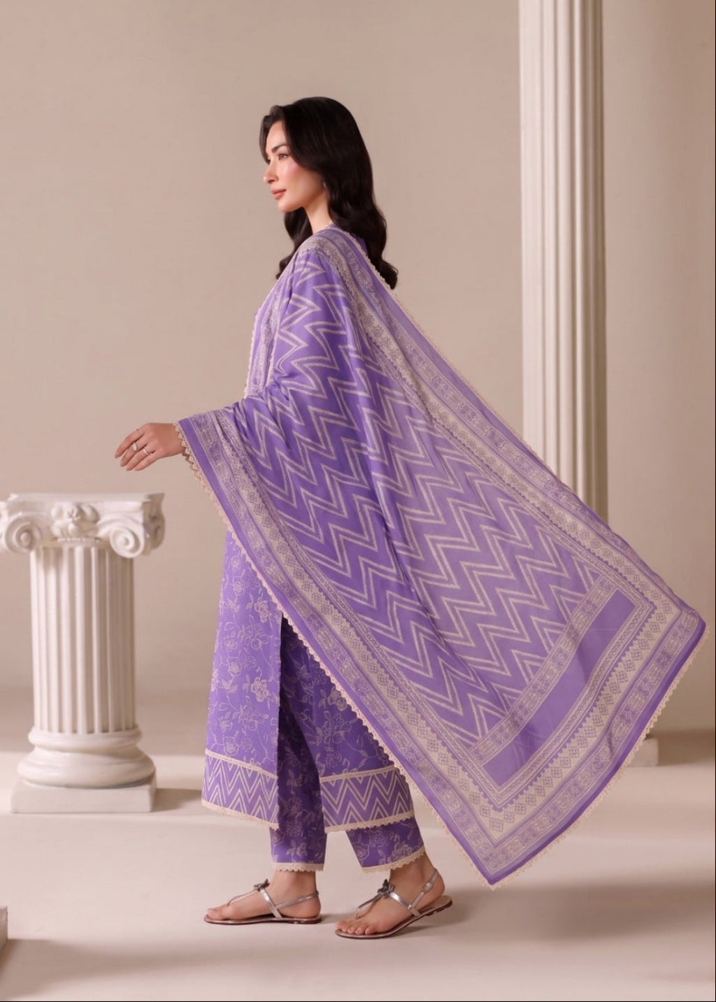 A woman models a purple three-piece traditional outfit with a matching patterned dupatta, standing in a classical setting with beige walls and white columns.