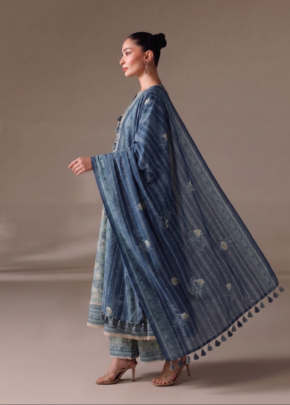 A woman models a traditional blue embroidered outfit with a matching dupatta and light blue patterned inner dress, photographed against a plain beige studio backdrop.