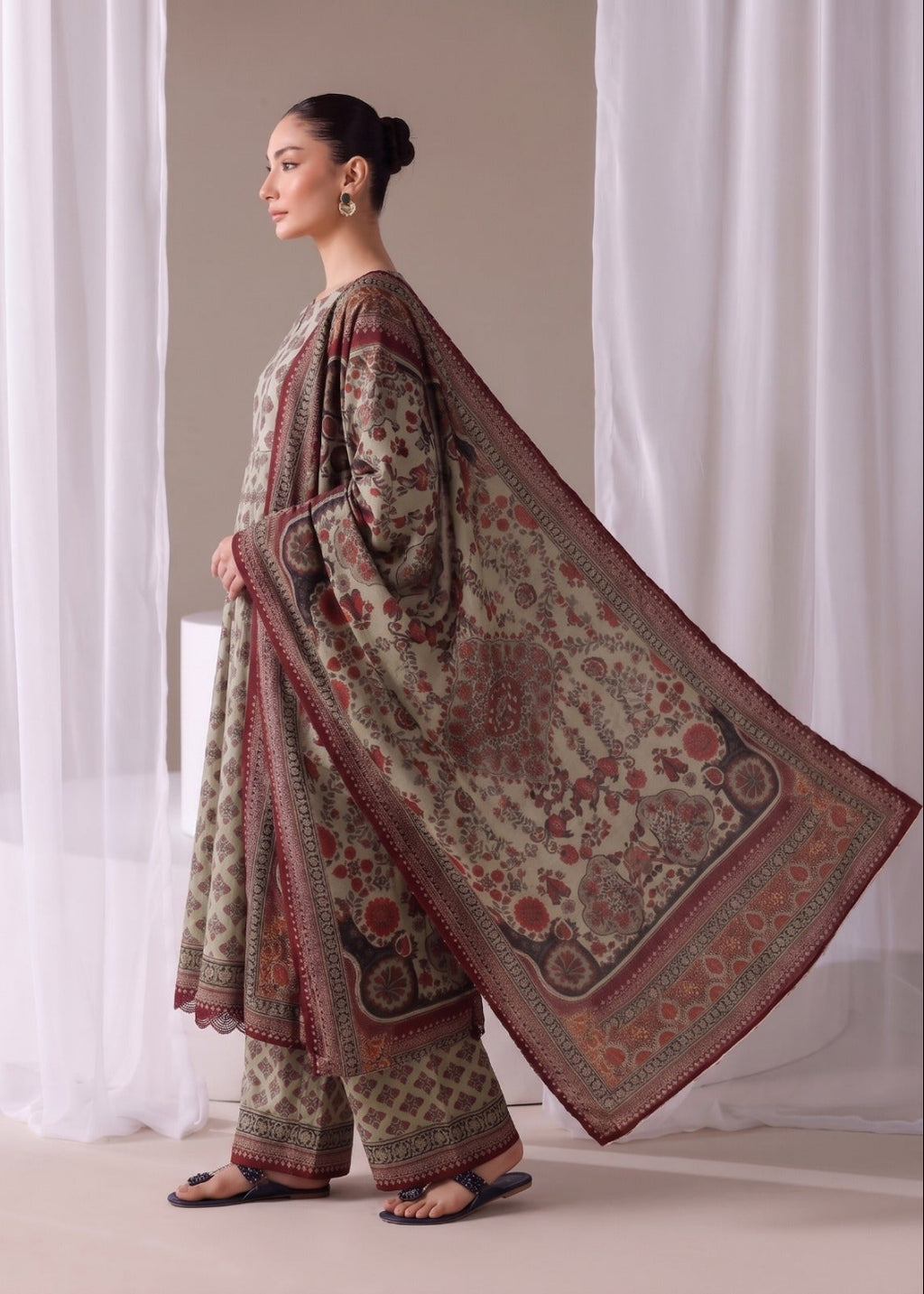 A woman modeling a traditional three-piece salwar kameez suit in a side profile view. The outfit features a beige kurta and matching trousers with intricate red and green floral patterns, paired with a large matching dupatta draped over her shoulder.