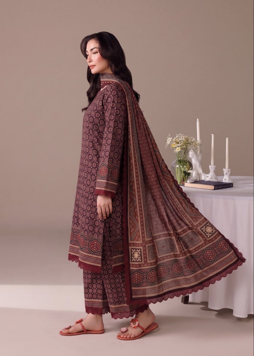 A woman modeling a traditional three-piece salwar kameez suit in maroon with intricate geometric patterns, paired with a matching dupatta, in a studio setting.