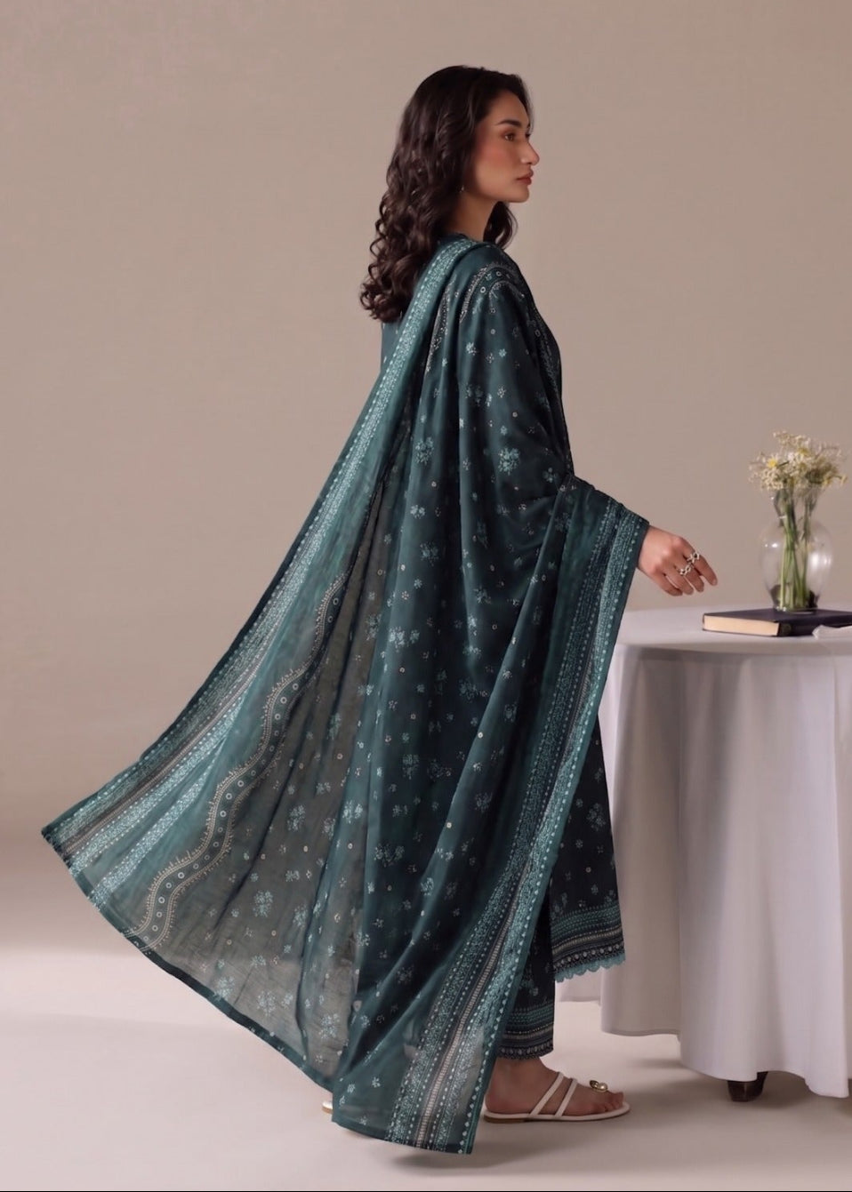 A woman wearing a dark teal green traditional outfit with a matching printed dupatta, standing in profile next to a white draped table with decorative items.