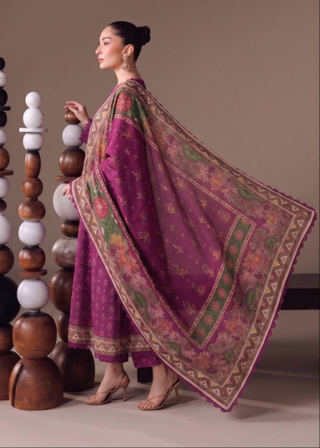 A woman models a traditional South Asian outfit featuring a magenta-purple kurta set with a matching printed dupatta, posed next to decorative wooden and white spheres on a neutral beige background.