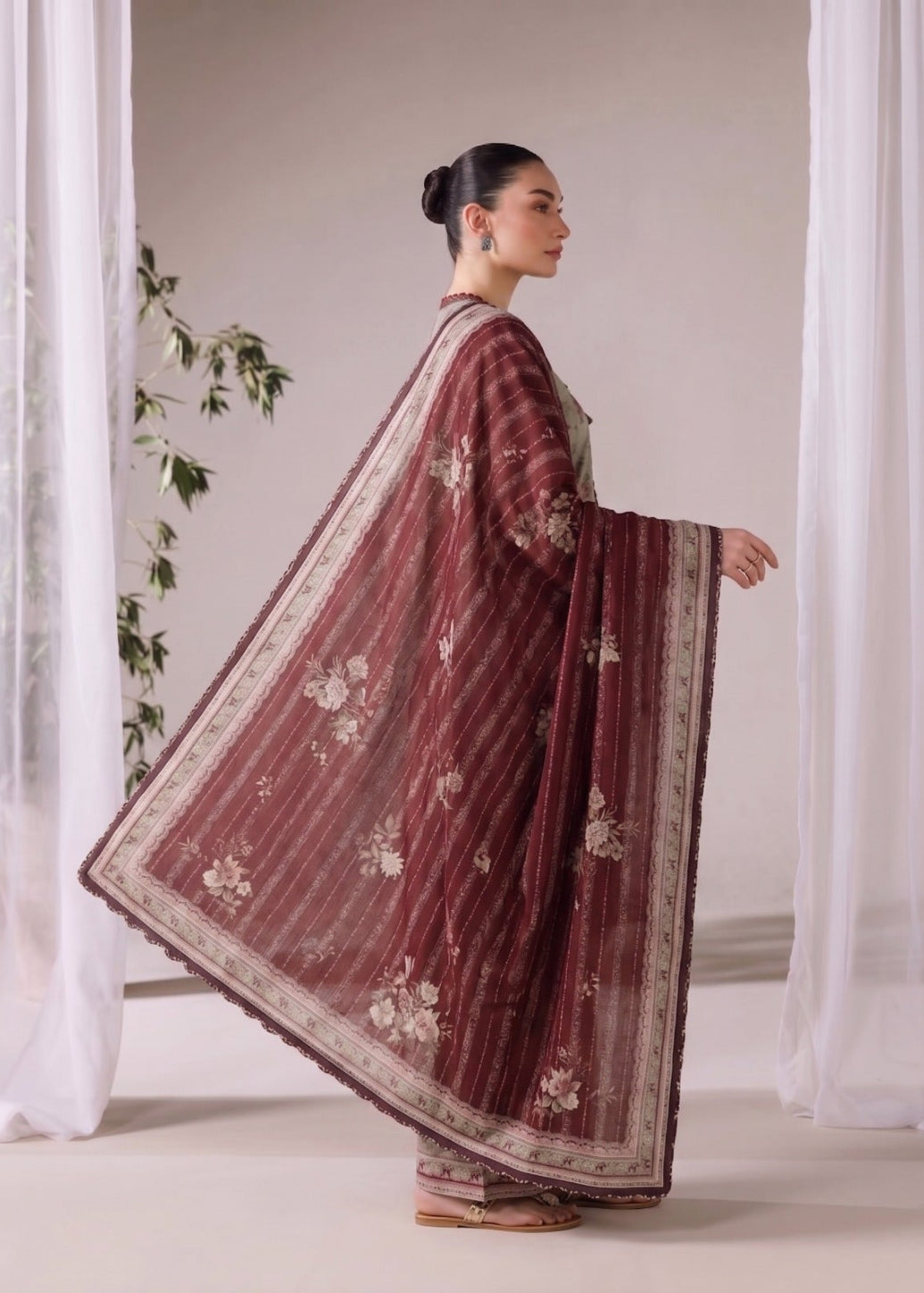 A woman in profile wearing a deep maroon embroidered dupatta and matching trousers, standing in a studio with white sheer curtains.