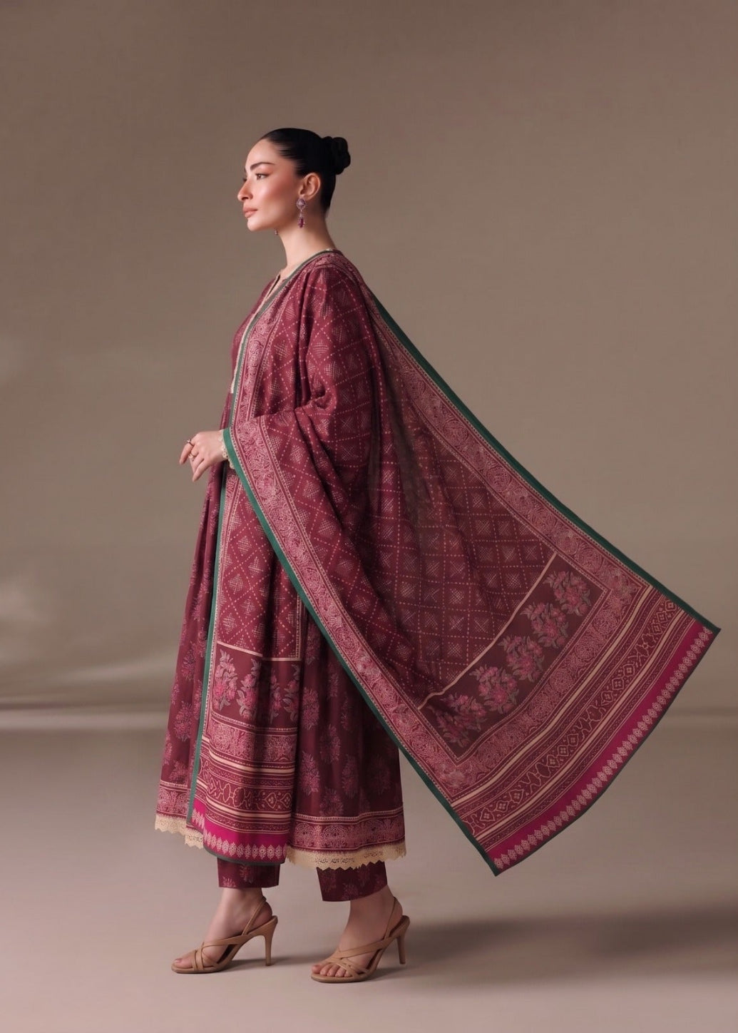 A woman modeling a traditional three-piece outfit in deep maroon with intricate gold and green printed patterns, featuring a matching dupatta, set against a neutral studio backdrop.