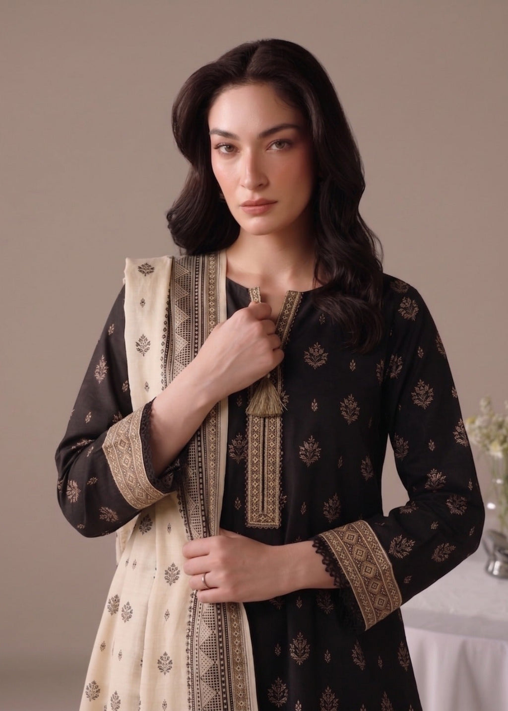 A woman modeling a black three-piece salwar kameez suit with beige block prints and a matching beige dupatta, set against a neutral beige background.