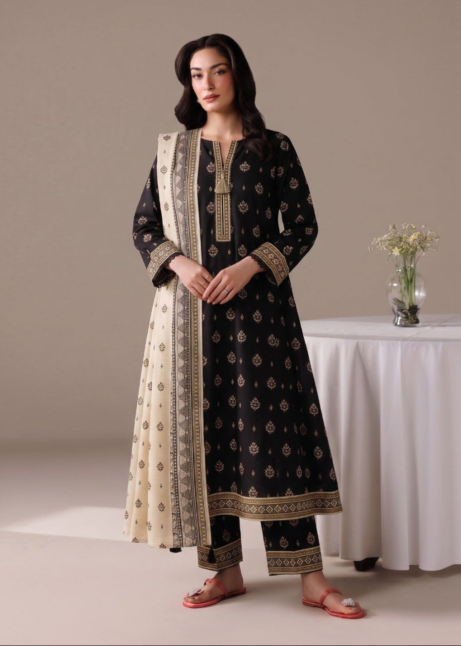 A woman models a black three-piece salwar kameez suit with beige and gold printed patterns, featuring a beige dupatta with a gold border, set against a neutral studio background with a white draped table.