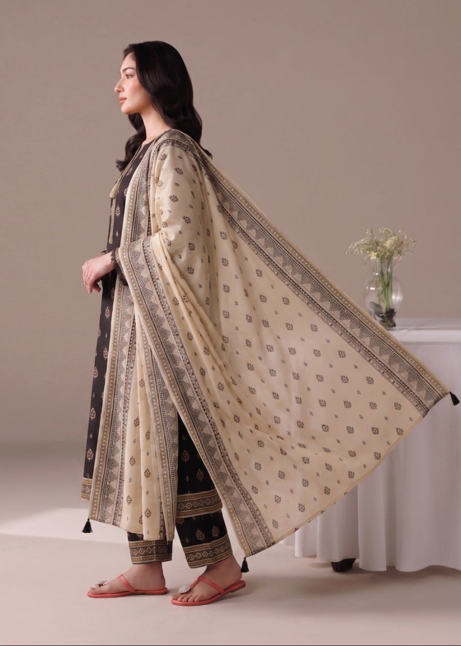 A woman stands in profile wearing a traditional South Asian outfit consisting of a beige kurta with brown floral motifs and a matching dupatta, paired with dark brown trousers. She is posed in front of a neutral beige studio backdrop with a white draped table and vase in the background.