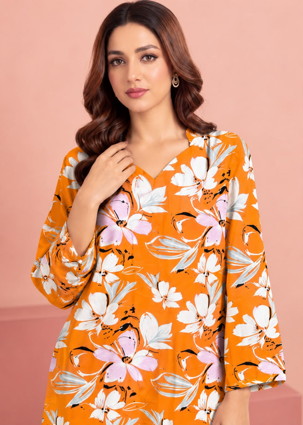 A woman wearing a bright orange V-neck blouse with a white, pink, and gray floral print, posed against a solid pink background.