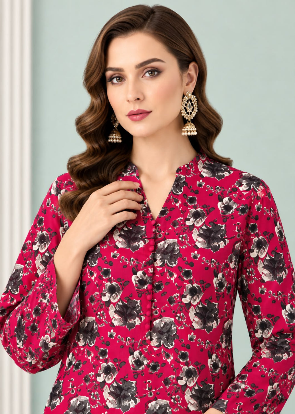 A woman wearing a bright pink long-sleeve blouse with a black and gray floral print, styled with ornate gold jhumka earrings against a pale green background.