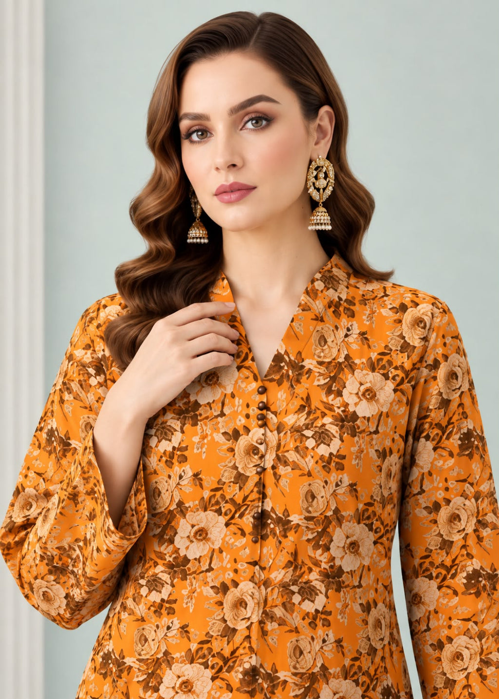 A woman wearing a mustard yellow floral printed long-sleeve kurta with brown and beige tones, paired with ornate gold-toned earrings, set against a soft light blue-gray background.