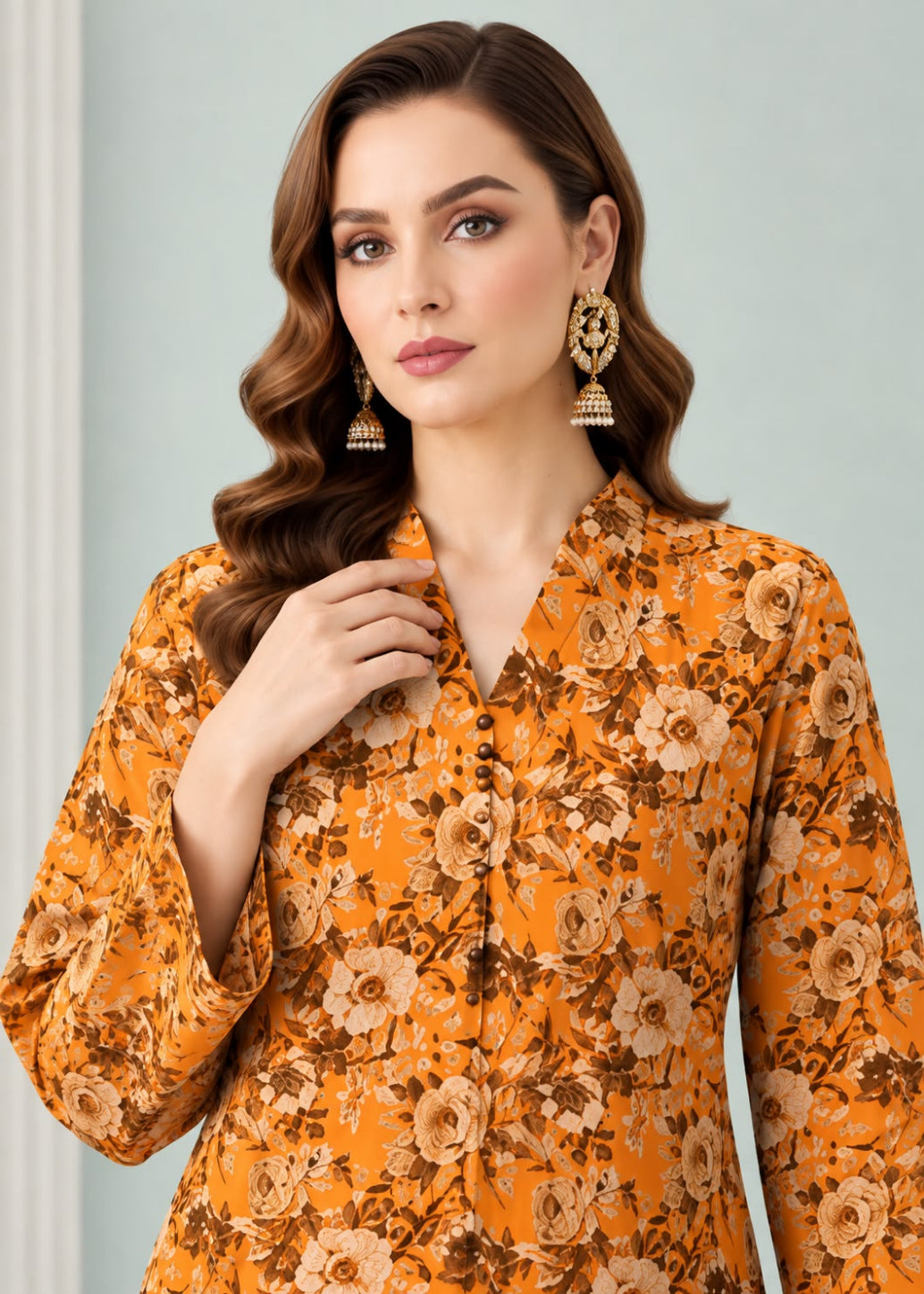A woman wearing a mustard yellow floral printed long-sleeve kurta with brown and beige tones, paired with ornate gold-toned earrings, set against a soft light blue-gray background.