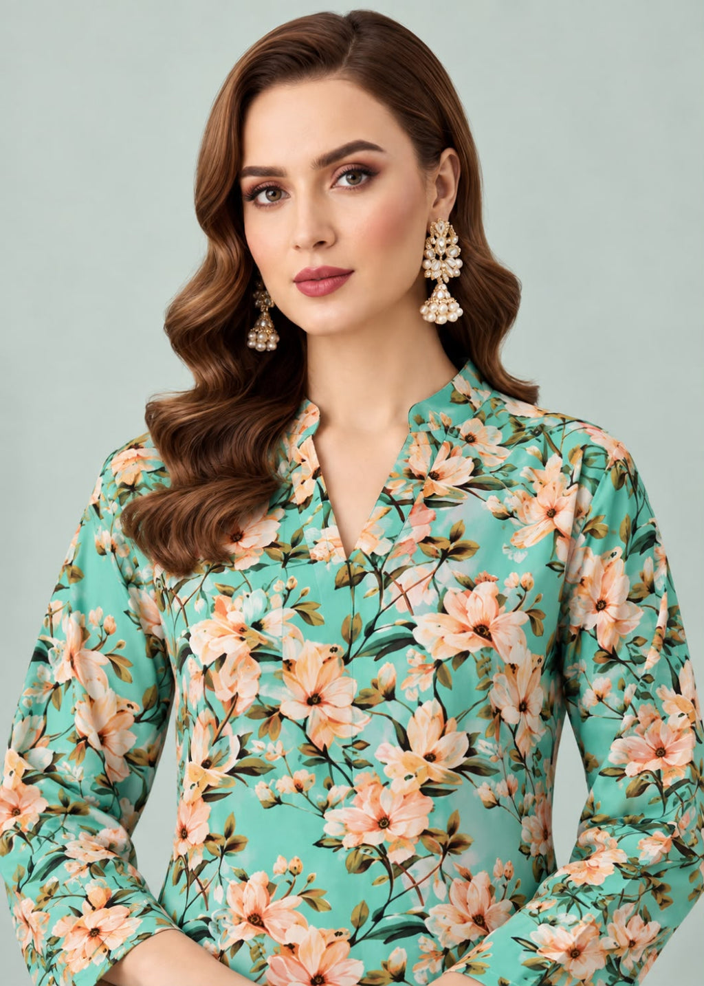 A woman wearing a mint green long-sleeve blouse with a peach and beige floral print, styled with statement earrings against a solid light gray background.