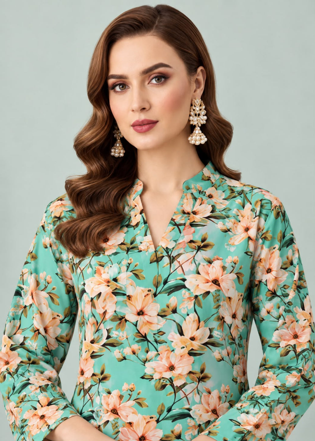 A woman wearing a mint green long-sleeve blouse with a peach and beige floral print, styled with statement earrings against a solid light gray background.