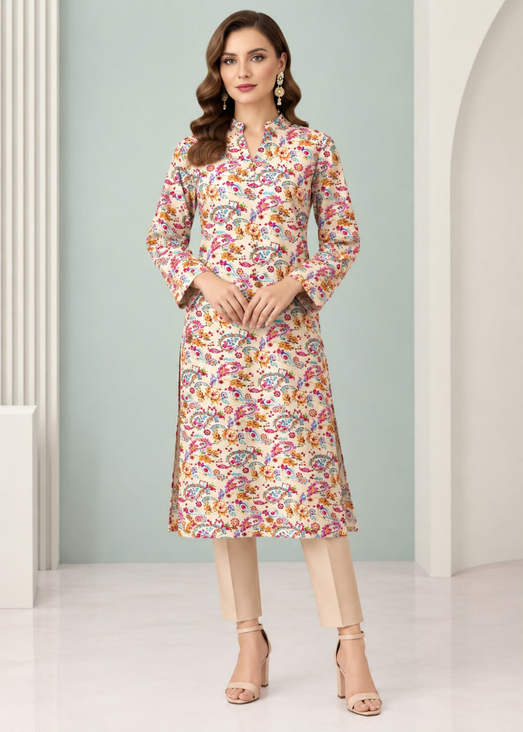 A woman models a long-sleeved, knee-length kurta with a dense multicolor floral and paisley print on a light beige base, paired with beige trousers and nude high-heeled sandals in a bright, modern studio setting.