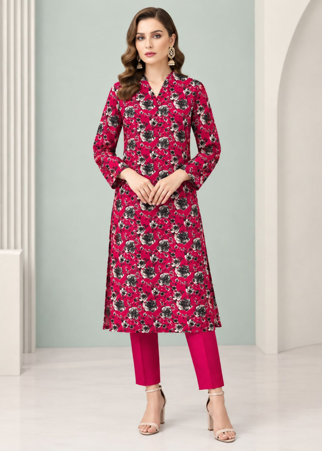 A woman modeling a long-sleeve floral printed kurta in magenta pink with black and white floral motifs, paired with matching pink pants and beige high-heeled sandals, set against a minimalist studio backdrop with white columns and arches.