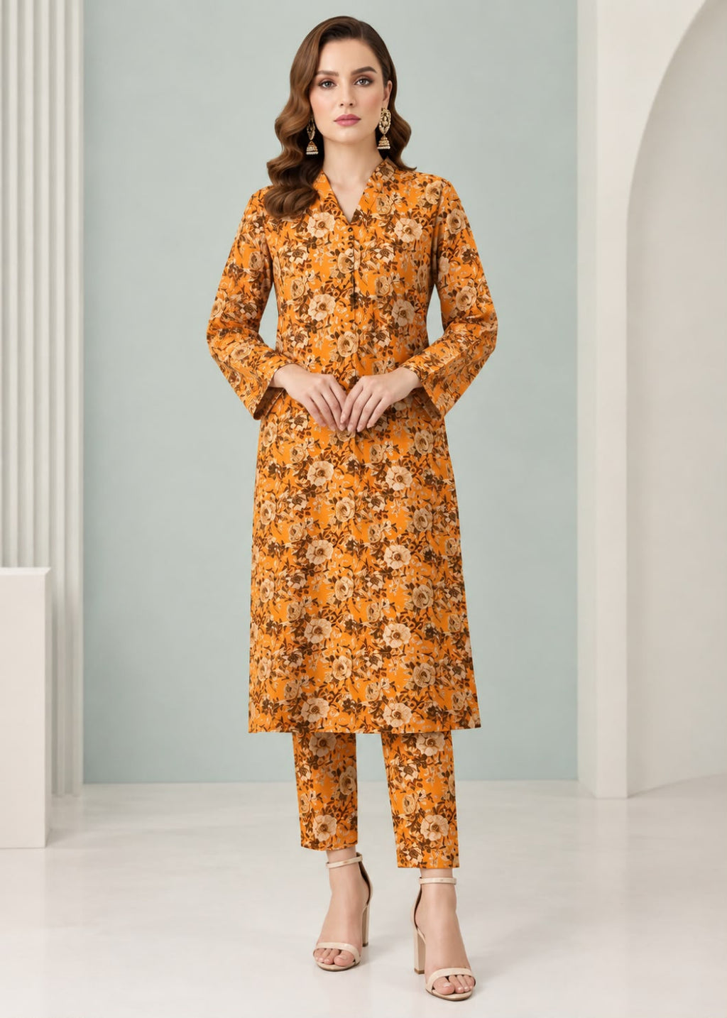 A woman modeling a mustard yellow and orange floral print kurta set with matching pants, standing in a studio with a light blue-gray and white background.