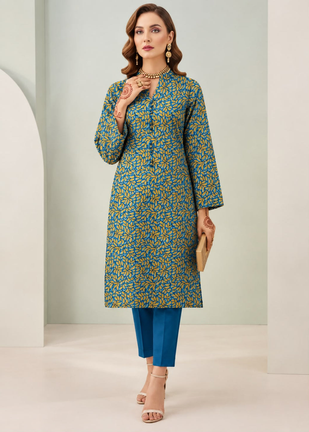A woman models a long-sleeved kurta with a blue and mustard yellow floral pattern, paired with blue pants and beige heels, standing in a minimalist studio setting.