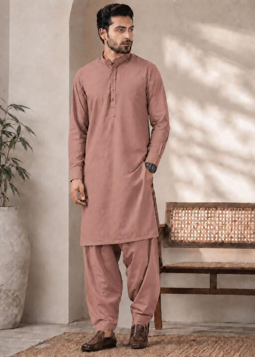 A man modeling a dusty rose pink traditional three-piece kurta set, consisting of a long-sleeve button-up tunic and matching loose-fitting trousers, standing in a styled indoor setting with a bench and potted plant.