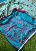 A folded and draped fabric set, likely a sari or scarf, featuring a turquoise blue base with intricate paisley and floral block prints in pink, red, and green, paired with a light mint green border section.