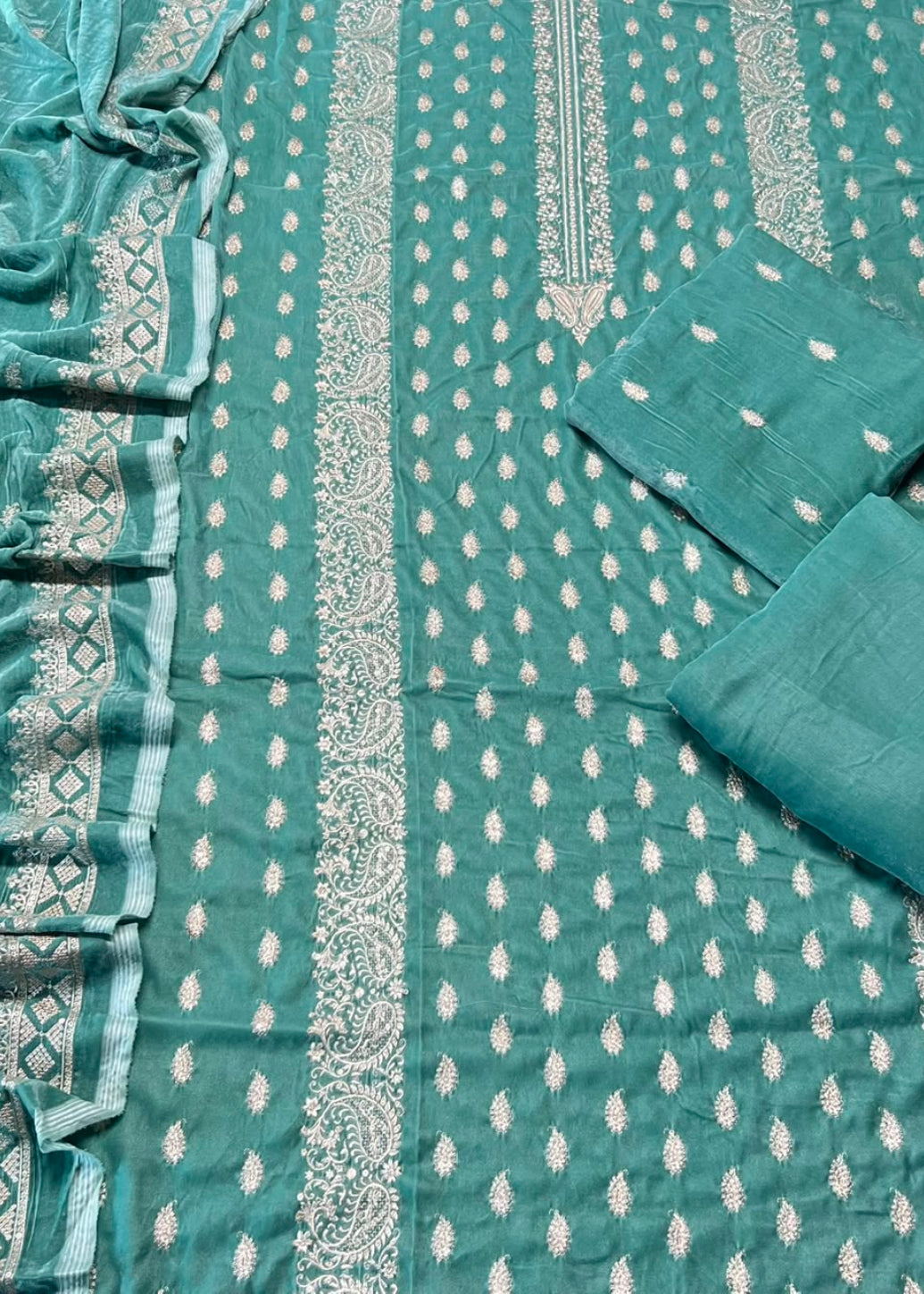 A teal green salwar suit fabric set laid flat, featuring a printed body fabric with vertical white and silver paisley borders and small motifs, matching folded fabric pieces, and a draped dupatta with ornate border patterns.