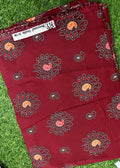 A folded piece of deep maroon cotton fabric with a printed bandhani style floral and paisley pattern in black, white, and orange, displayed on green grass.