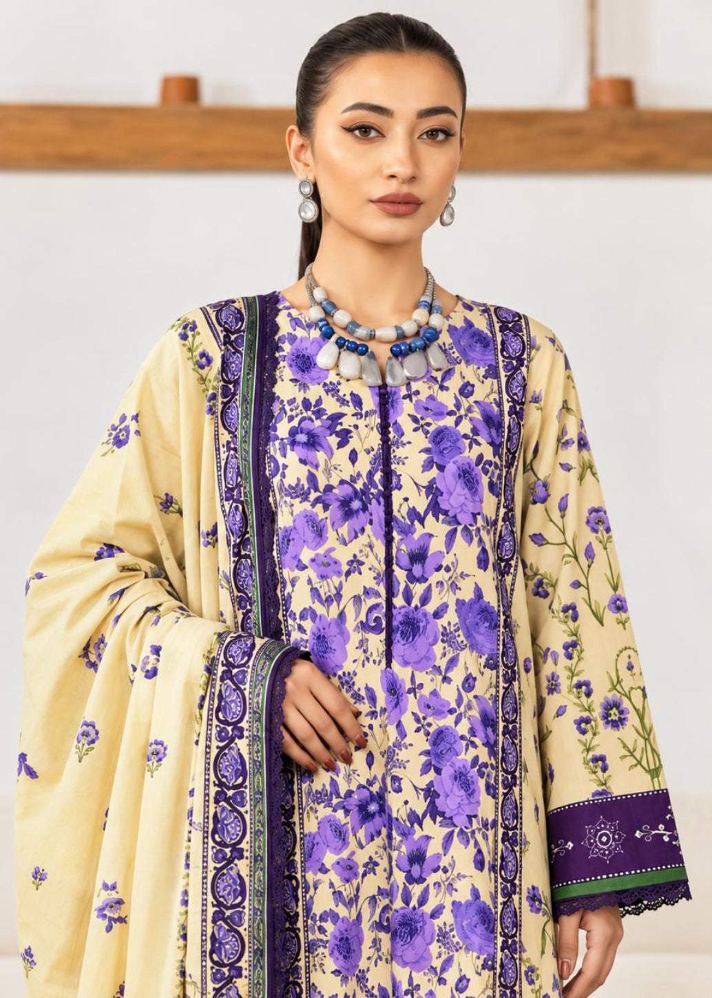 A woman modeling a traditional three-piece salwar kameez suit in beige and purple. The kurta features a vibrant purple floral print on a beige base, with matching purple and green patterned borders.