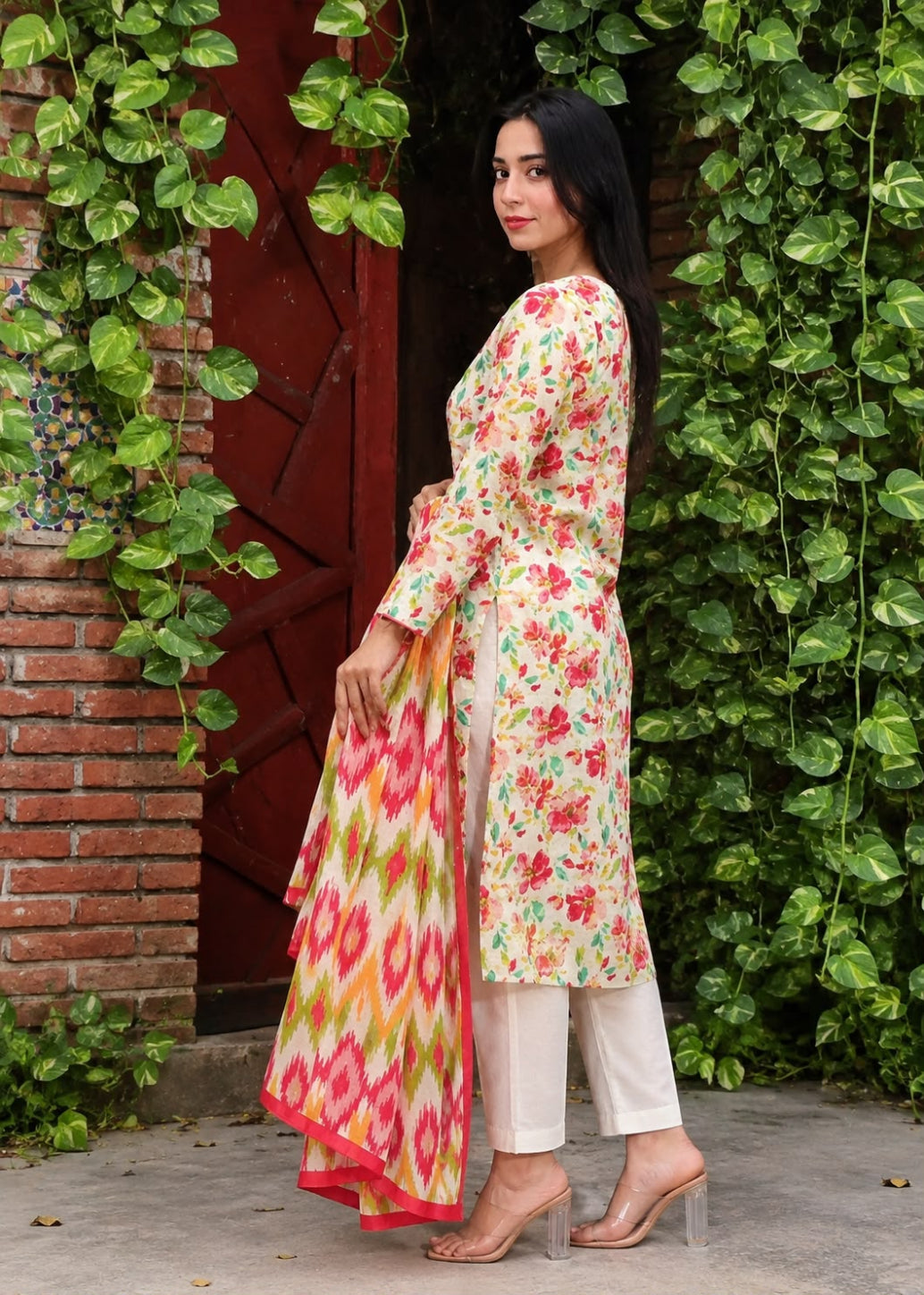 A woman stands outdoors in front of a brick wall and green ivy, wearing a floral long kurta with a matching printed dupatta and white pants.