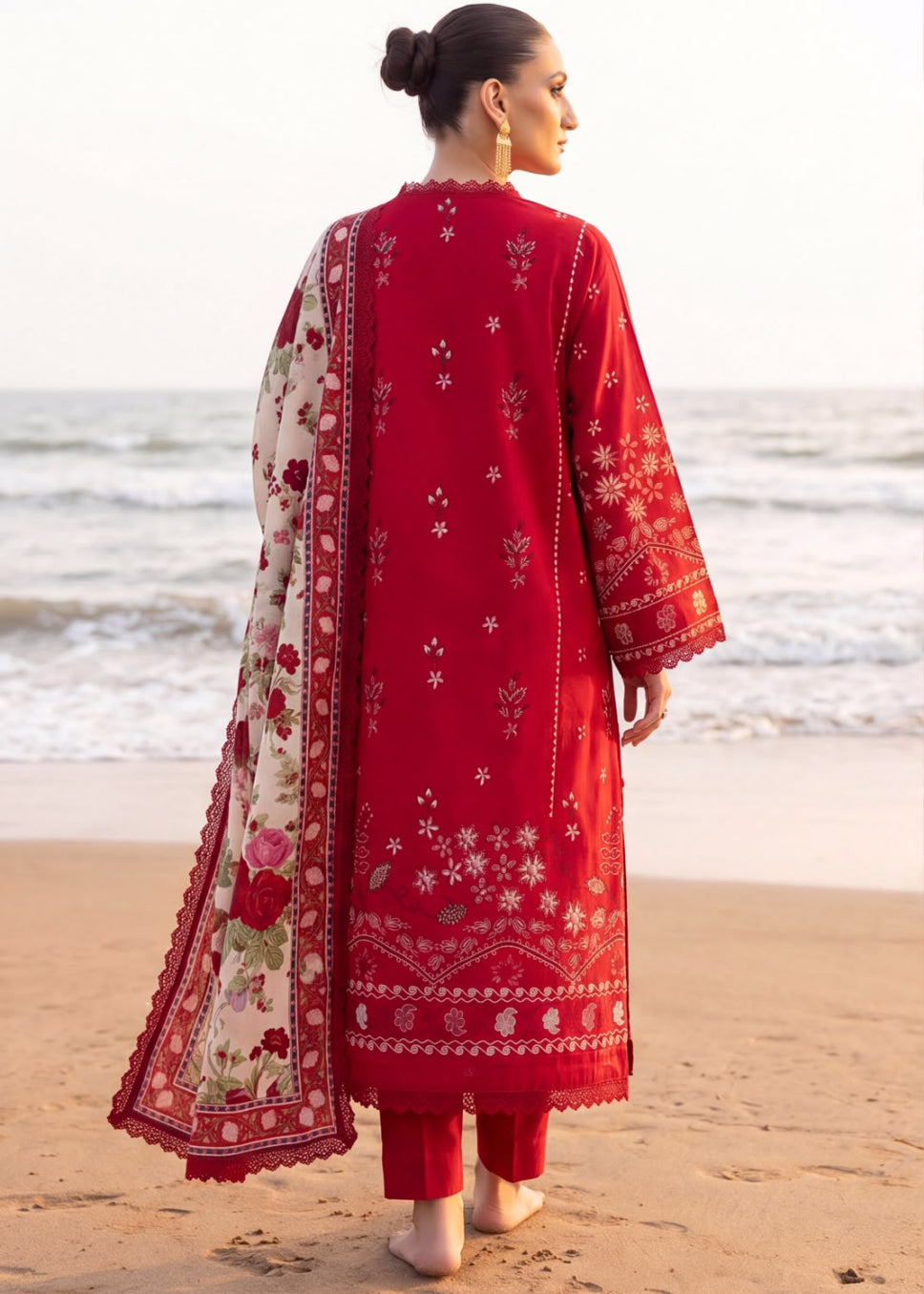 A woman stands on a sandy beach facing the ocean, wearing a red traditional outfit with white floral embroidery and a matching red dupatta with a cream floral border.