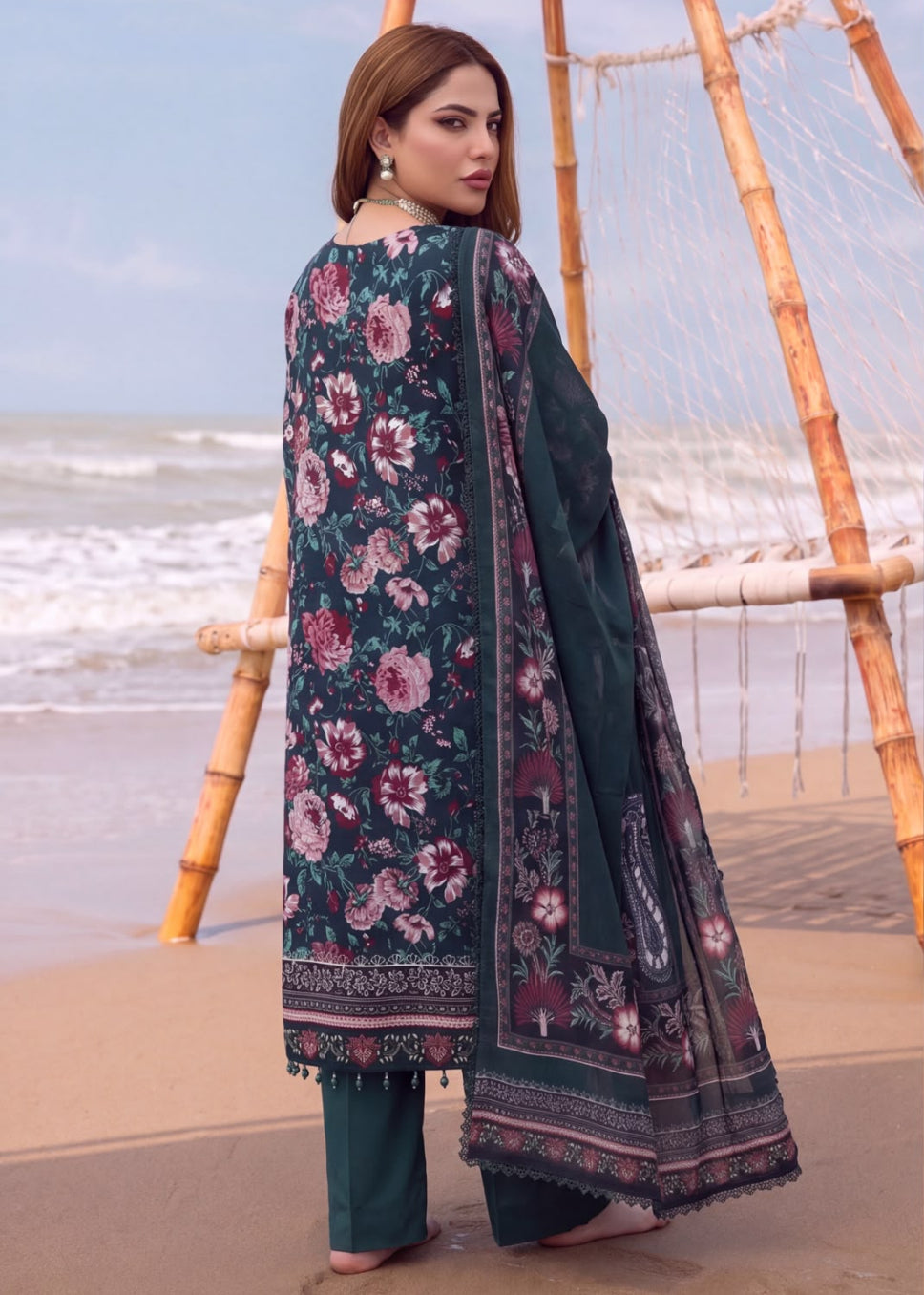 A woman stands on a sandy beach wearing a dark teal floral salwar kameez suit with wide-leg trousers and a matching dupatta. The outfit features pink and purple floral patterns on a dark teal base with ornate borders.