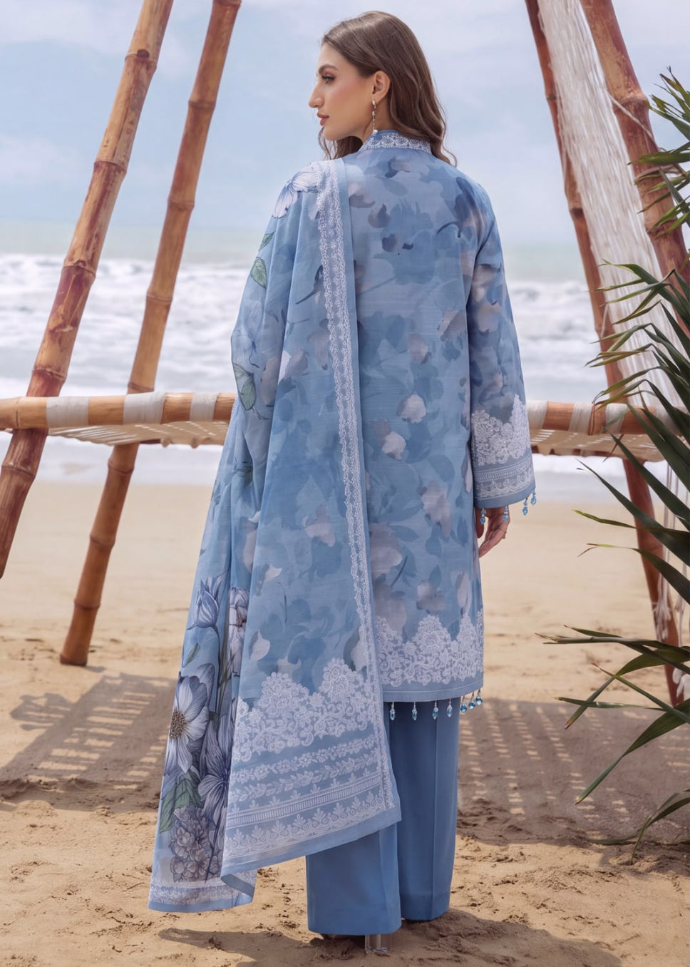 A woman stands on a sandy beach wearing a light blue printed traditional outfit with a matching dupatta and wide-leg trousers. Bamboo arches and ocean waves are visible in the background.