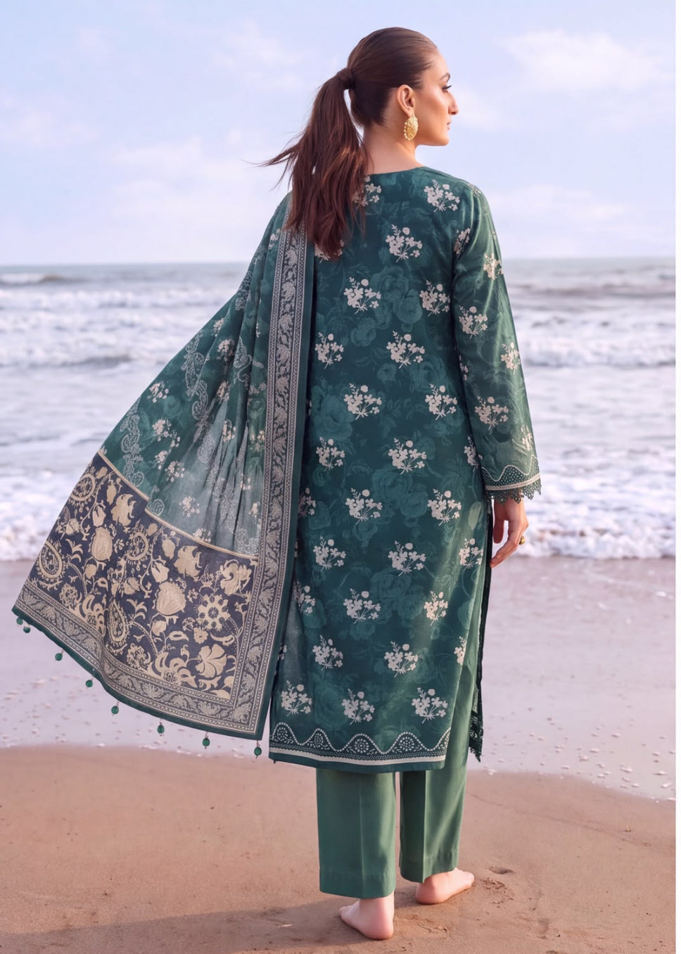 A woman stands on a sandy beach facing the ocean, wearing a dark green traditional outfit with white floral embroidery, matching pants, and a sheer embroidered dupatta.