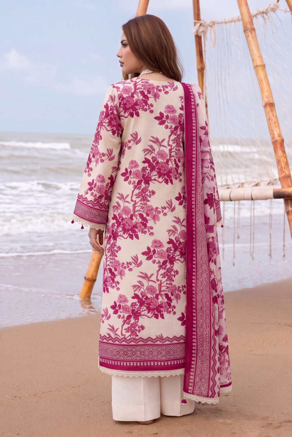 A woman stands on a sandy beach facing the ocean, wearing a cream-colored three-piece traditional outfit with a vibrant magenta floral print and matching dupatta.